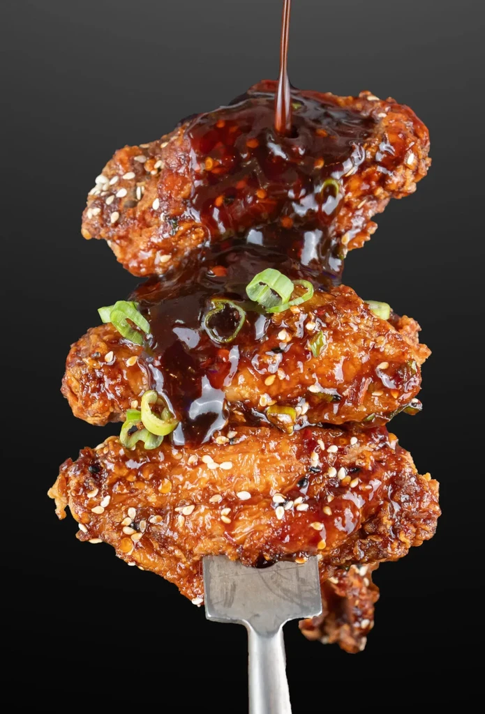 Crispy fried chicken wings stacked, dark sauce pouring, sesame and green onions.