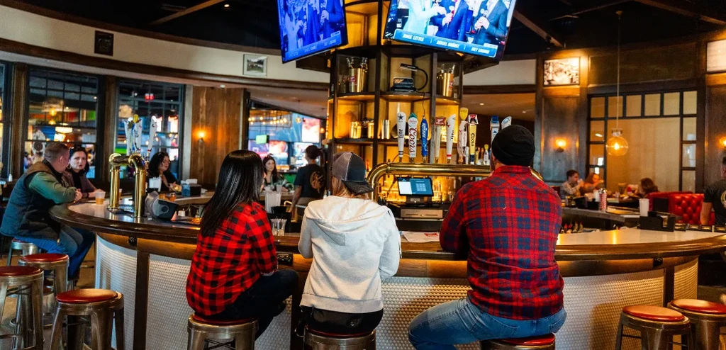 Patrons sit at a lively wooden bar with beer taps and sports TVs.
