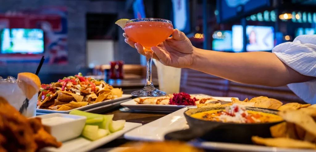 Hand holding orange cocktail, nachos, and dips in a bar setting.