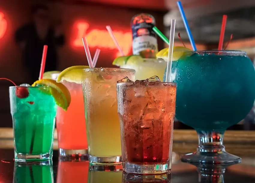 Five colorful cocktails with garnishes on a bar, neon background.