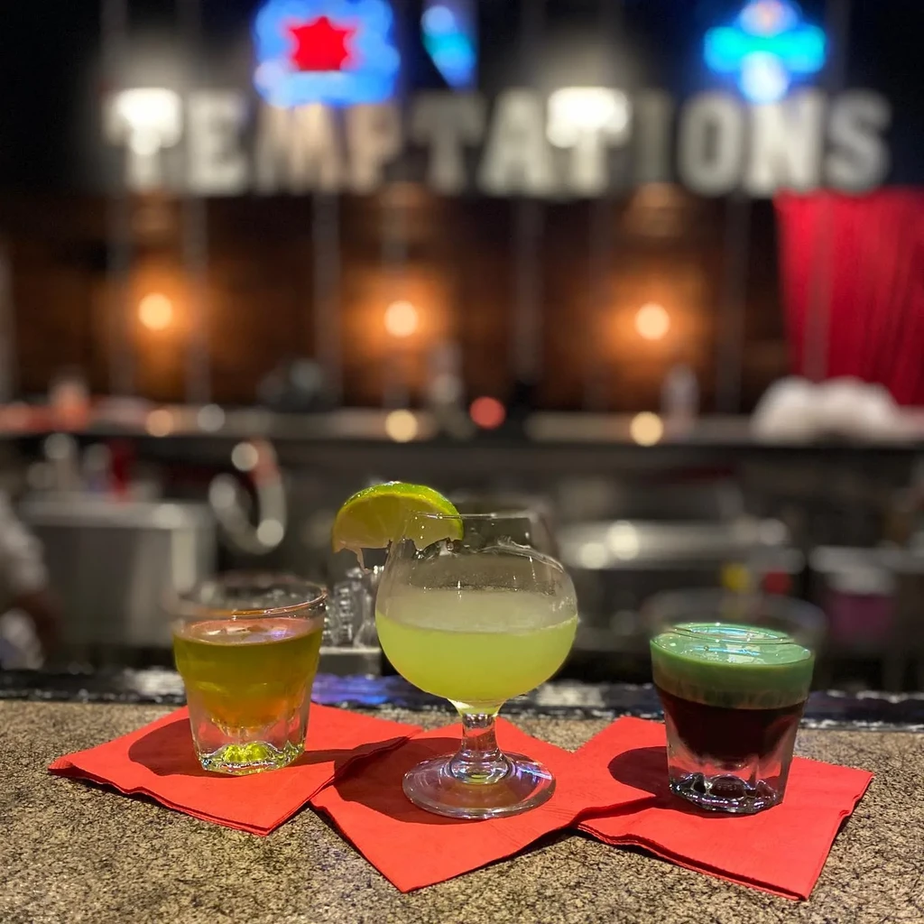 Three drinks on a bar counter with a blurred "TEMPTATIONS" sign.