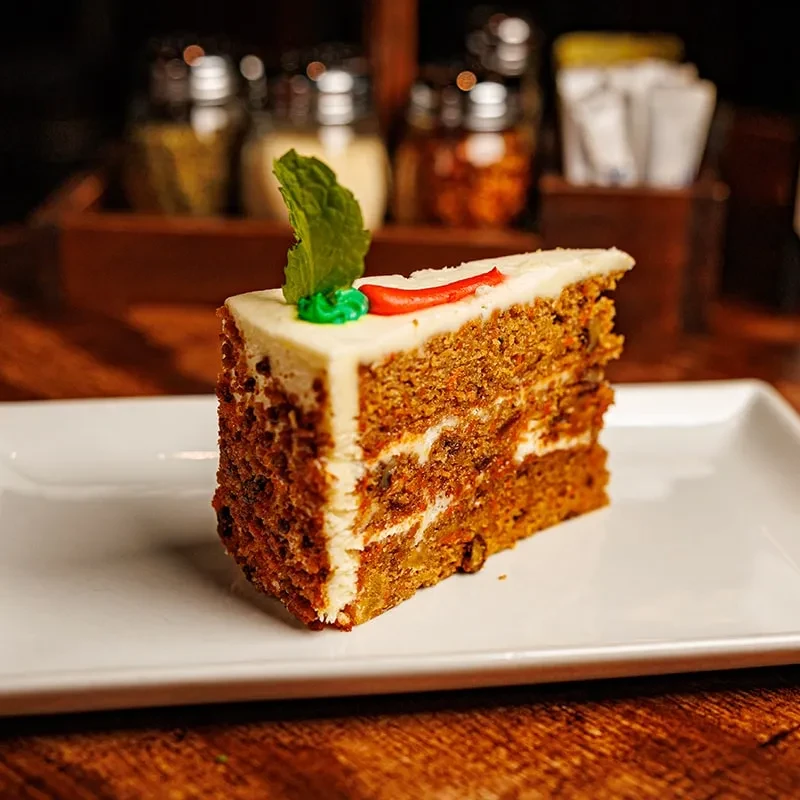 Slice of carrot cake with white frosting, mint leaf, and red carrot.
