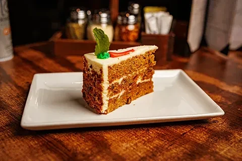 Slice of carrot cake with cream cheese frosting, mint leaf, and red garnish.