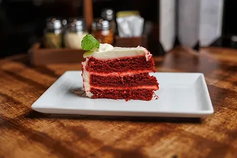 Red velvet cake slice with thick white frosting and mint leaf on white plate.