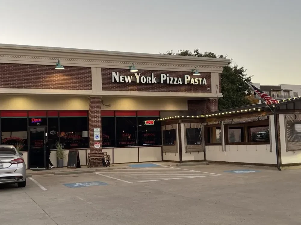 Illuminated 'New York Pizza Pasta' restaurant exterior at dusk with patio.
