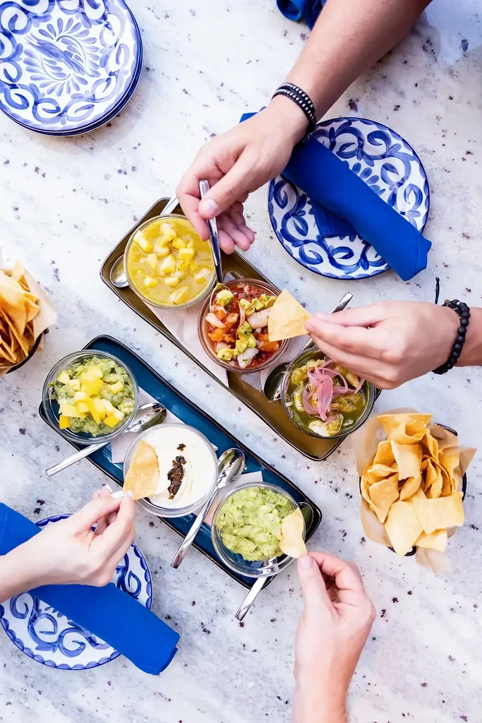 Overhead table view: colorful dips, ceviches, chips, and hands sharing a meal.