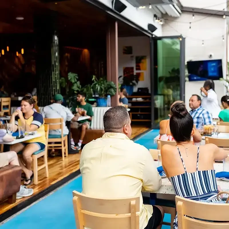 Bustling indoor-outdoor restaurant with diners, bright blue floor, and string lights.
