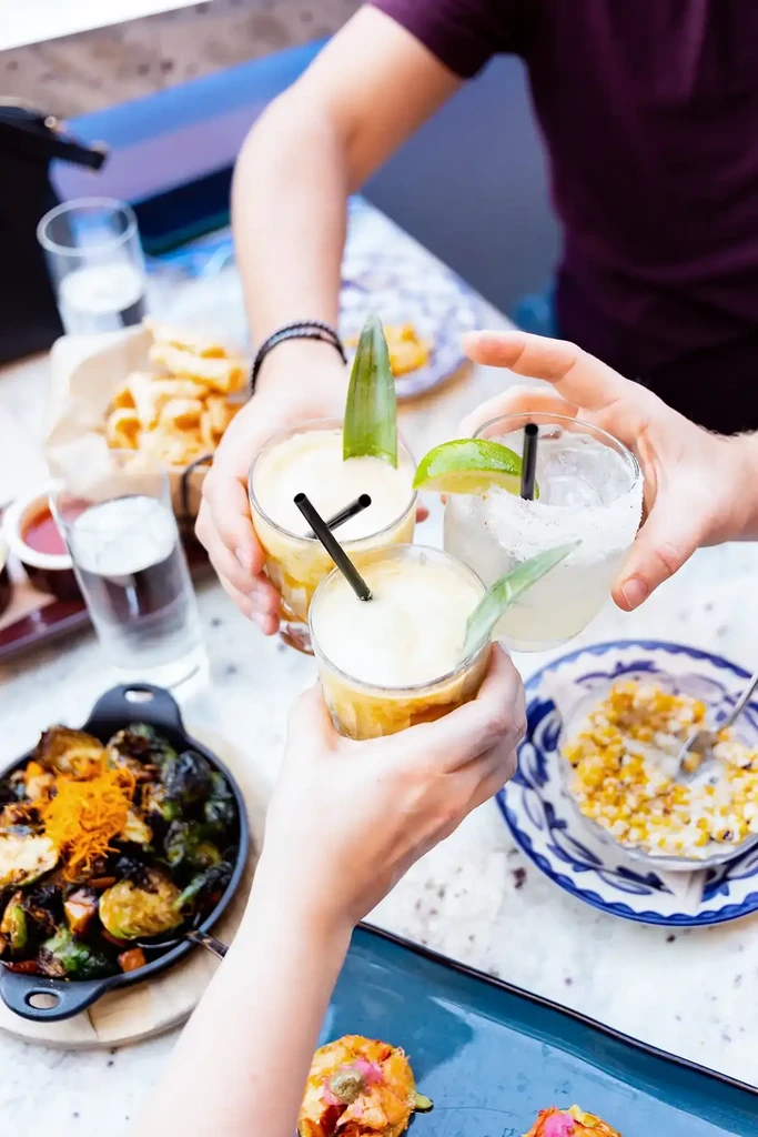 Overhead view: Three hands toasting drinks over varied food.