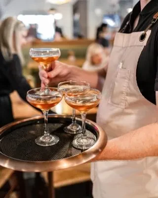 Server's hands holding a tray of amber cocktails in coupe glasses.