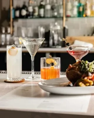 Four garnished cocktails and a plated dish on a bar.