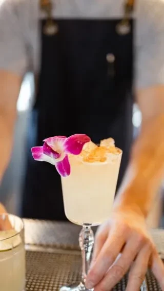 Elegant light yellow cocktail with purple orchid, blurred bartender in background.