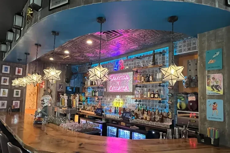 Vibrant bar with long counter, neon signs, skull figure, and Lotería art.