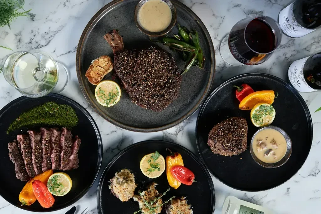 Overhead flat lay of gourmet steak dishes, roasted vegetables, sauces, and wine.