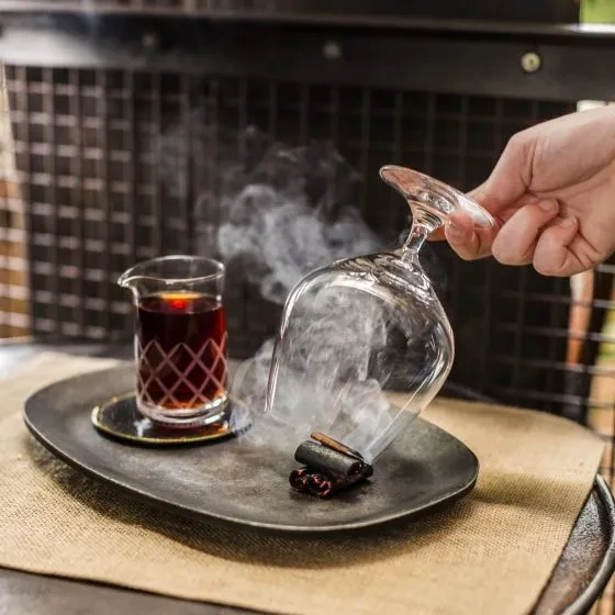 Hand holds inverted glass trapping smoke over spices; pitcher of drink nearby.