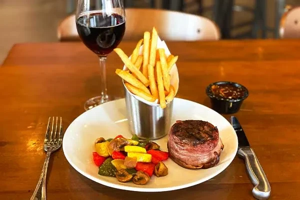 Seared steak, colorful vegetables, fries, and red wine on a wooden table.