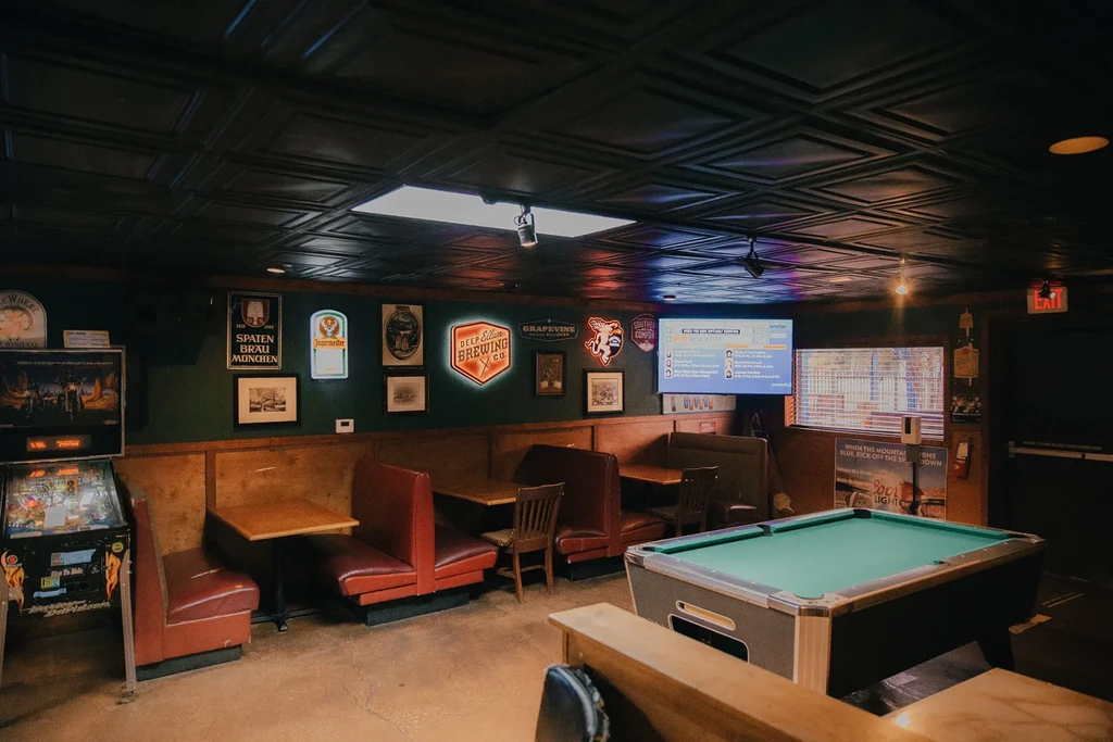 A dimly lit bar interior with a pool table, red booths, and neon signs.