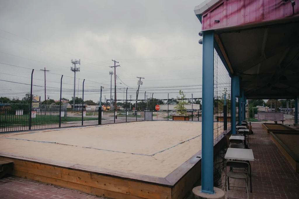 Empty sand volleyball court, covered patio, background utility poles, overcast sky.