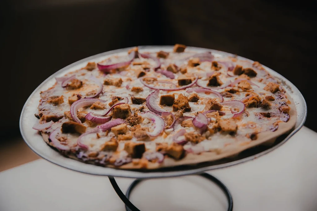 Pizza topped with cheese, cubed protein, and red onion on a stand.