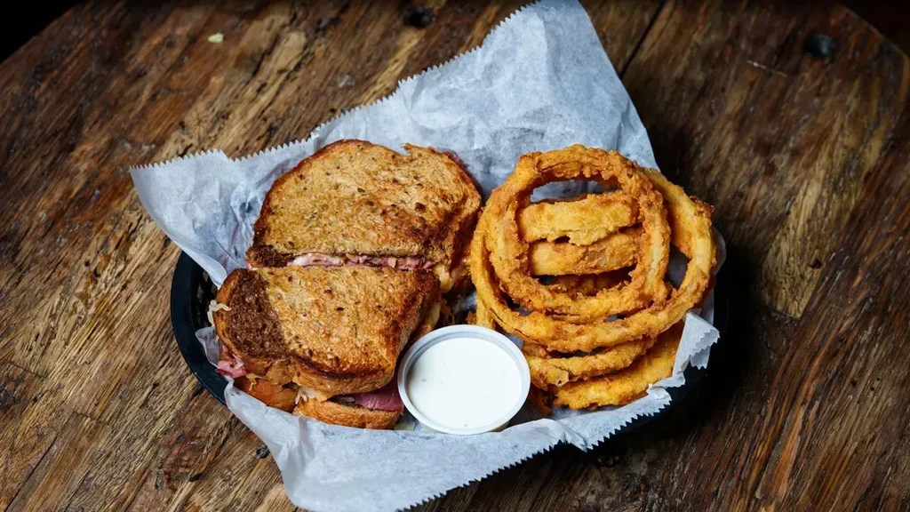 Close-up of a sandwich, onion rings, and creamy dip in a basket.
