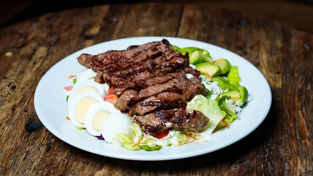 Grilled steak salad with eggs, avocado, blue cheese on white plate.