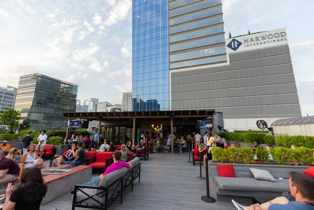 Rooftop bar with patrons, lounge seating, fire pits, and city view.