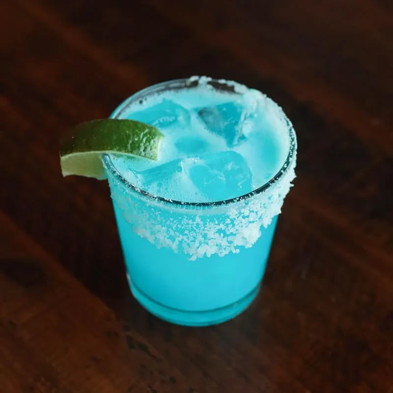 Blue cocktail with ice, lime, and frosted rim on a wooden surface.