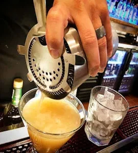 Bartender pours frothy orange drink from shaker into martini glass.