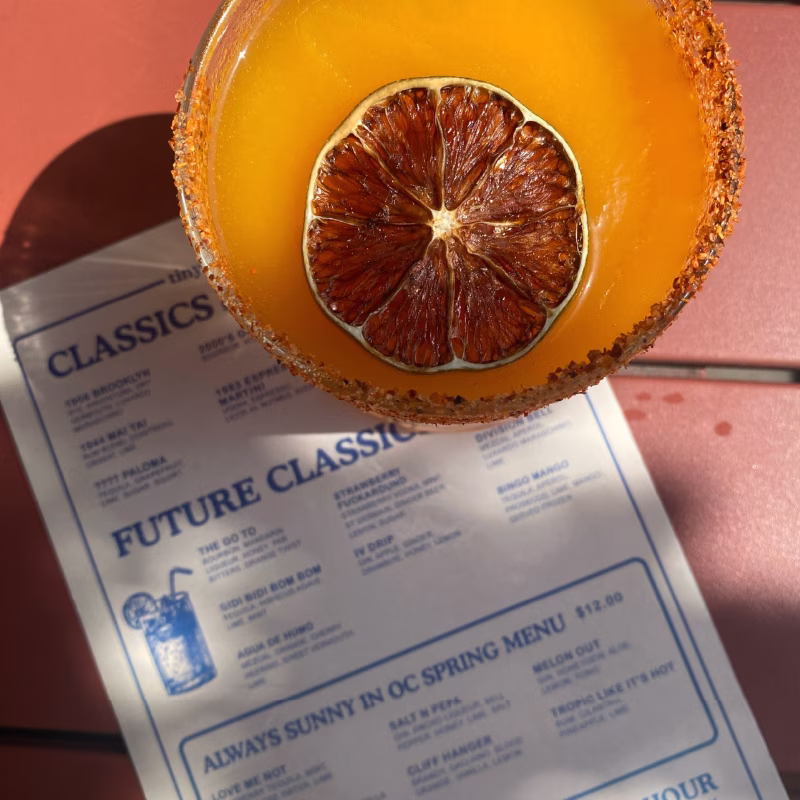 Spicy-rimmed orange cocktail with dried citrus slice and menu.