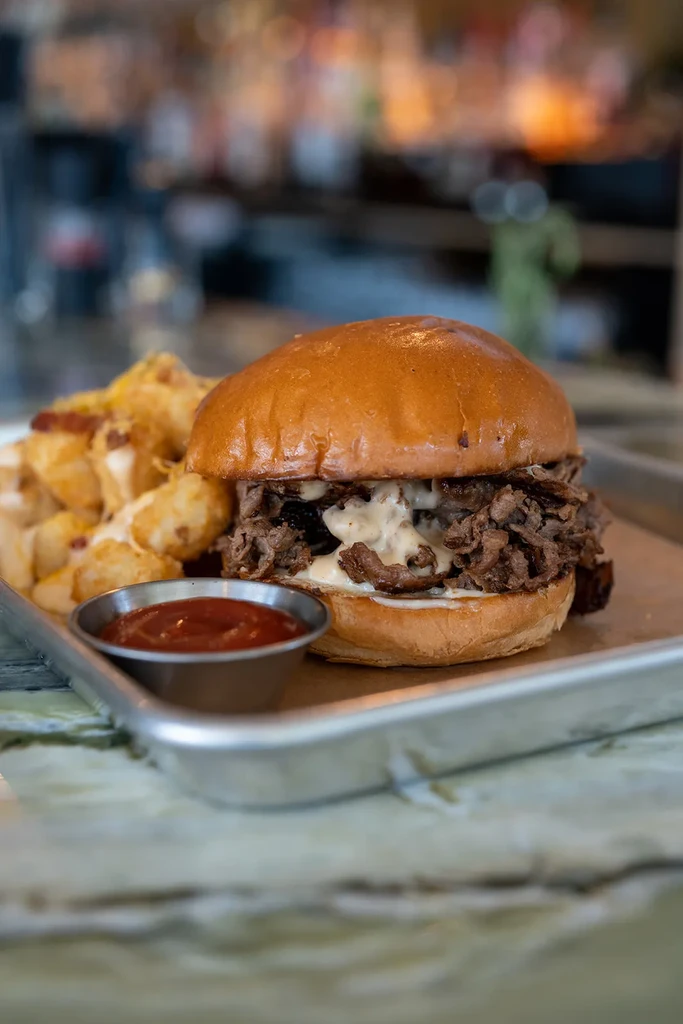 Close-up of a meat sandwich, tater tots, and red sauce on a tray.