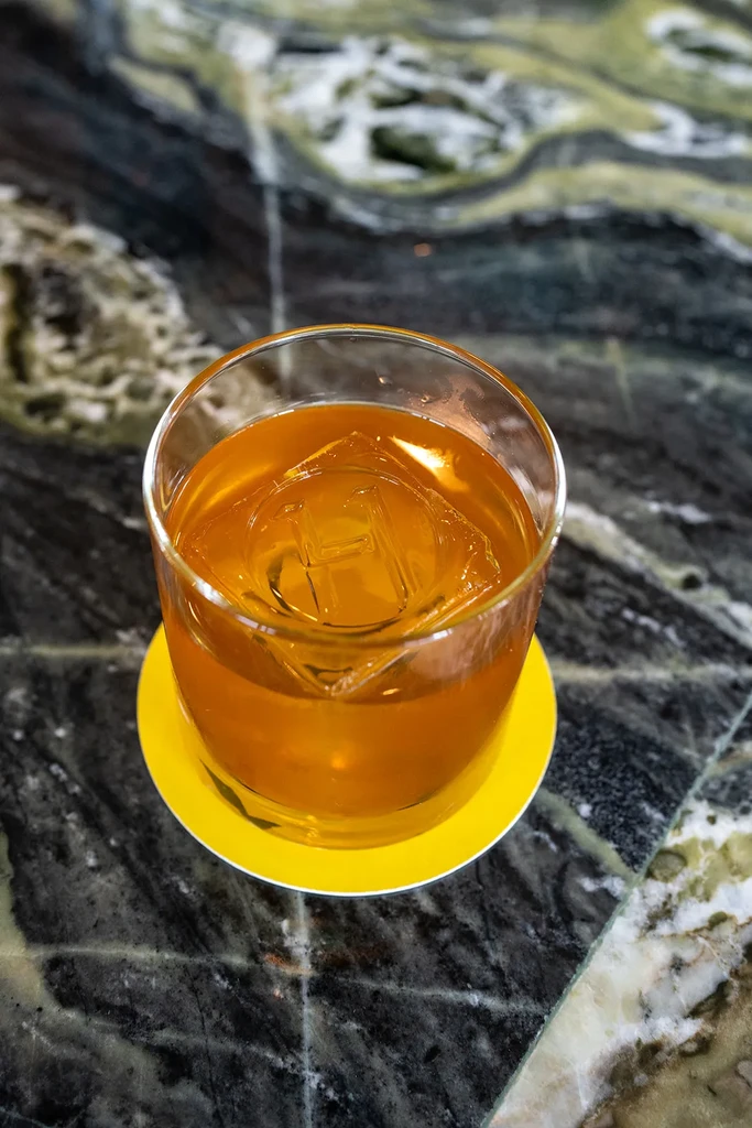 Amber drink with 'H' embossed ice cube on a yellow coaster.