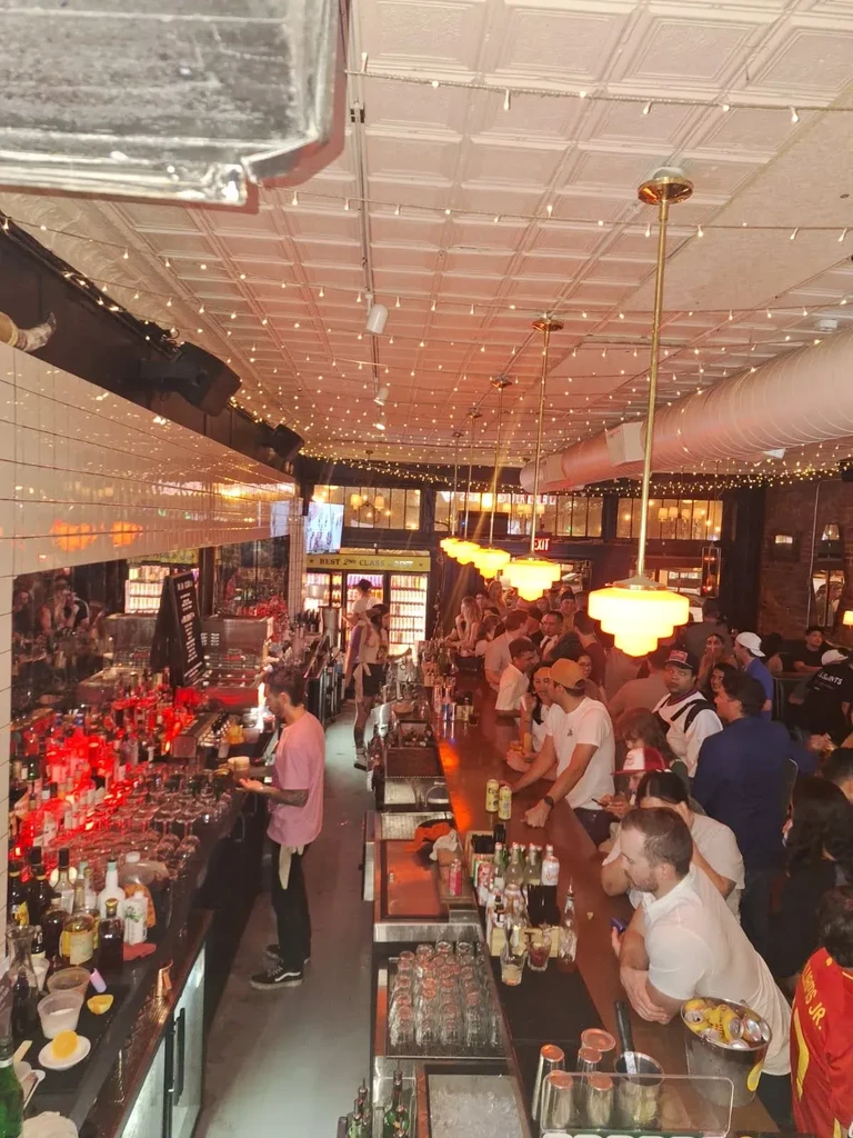 A crowded, lively bar interior with patrons, bartender, and colorful lights.