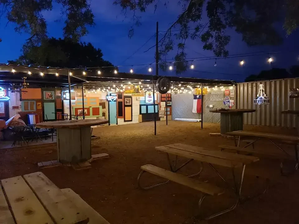 Illuminated outdoor beer garden at dusk with picnic tables and eclectic walls.