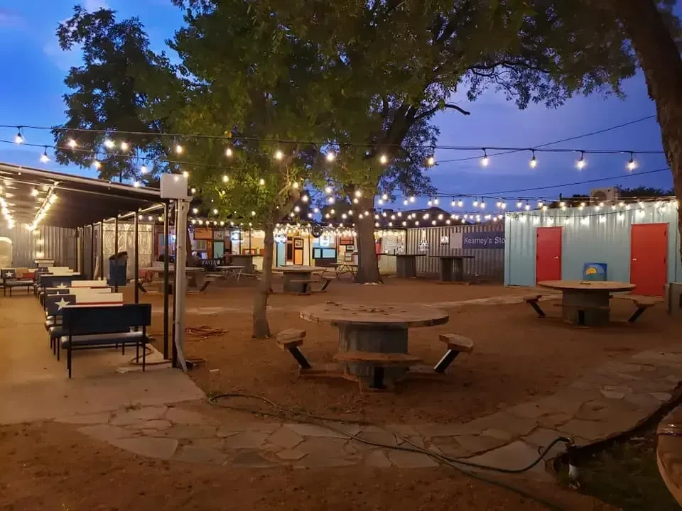 Outdoor eating area at dusk with string lights, picnic tables, shipping containers.