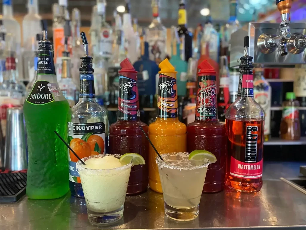 Two cocktails with salted rims and lime, liquor bottles behind.