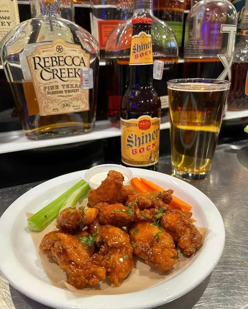 Glossy chicken wings, veggies, dip, whiskey, and beer on a metal bar.