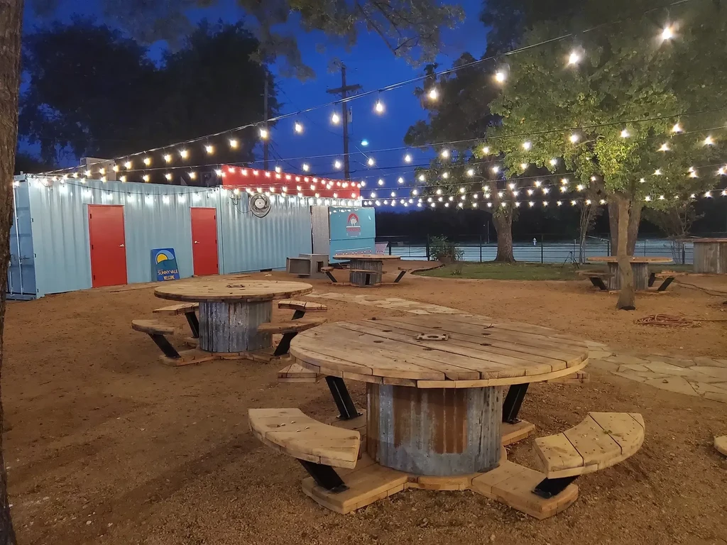 Outdoor dining with wooden spool tables, shipping containers, and string lights.
