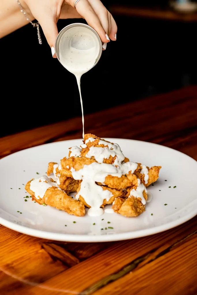 Hand pouring white sauce over crispy golden fried food.