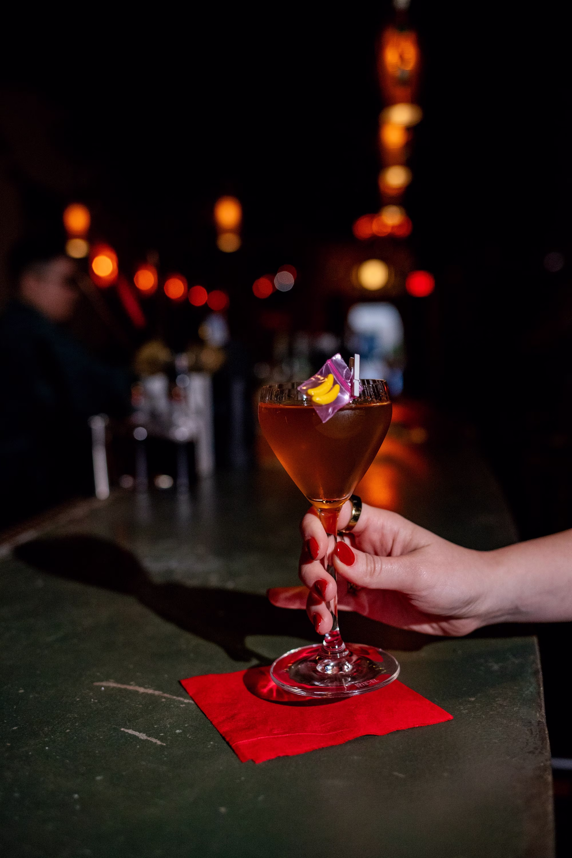 Hand holding a cocktail with banana candy garnish in a dark bar.