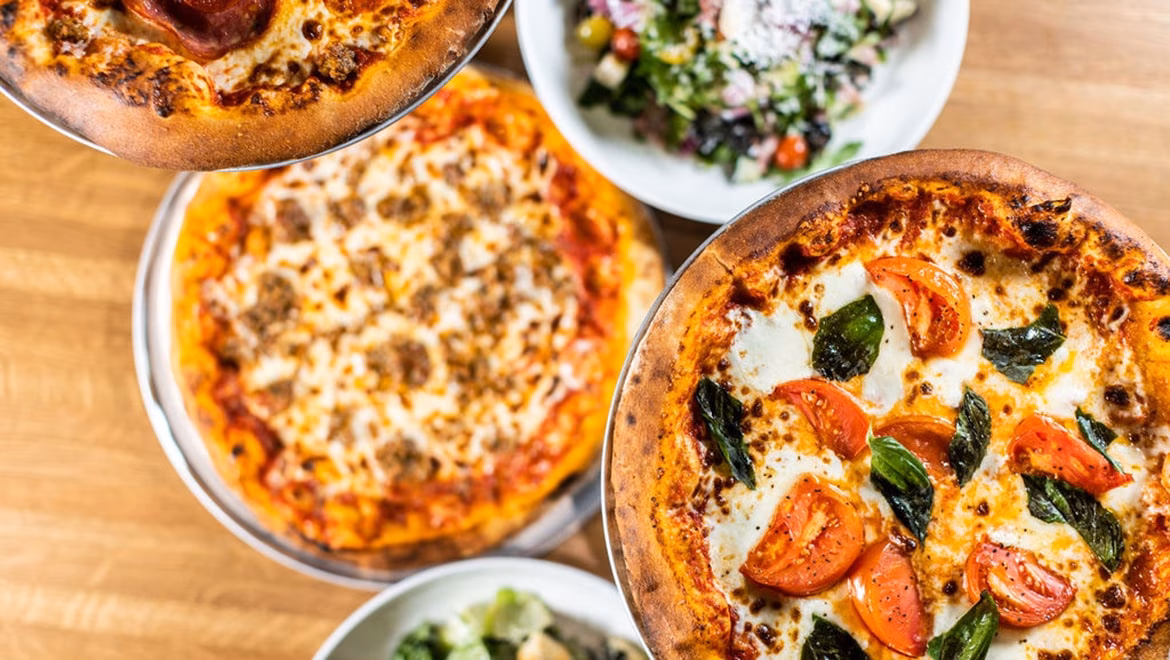 Three pizzas and two salads on a wooden table, overhead view.