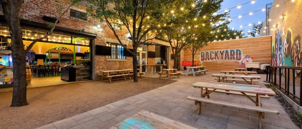 Empty 'BACKYARD' restaurant patio with string lights and picnic tables at dusk.