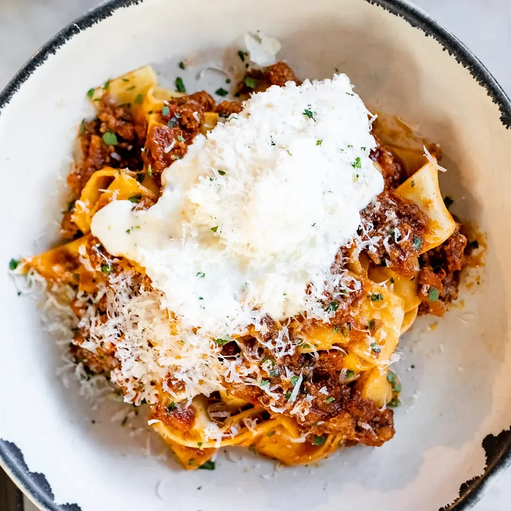 Pappardelle bolognese topped with burrata and grated parmesan in bowl