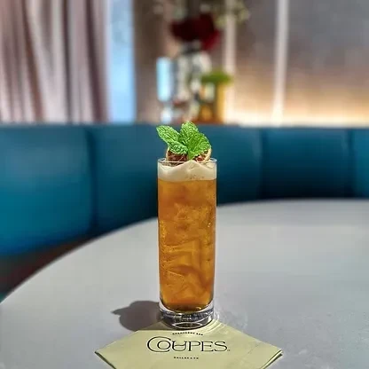 Amber drink with foam, mint, citrus, and a "COUPES" napkin.