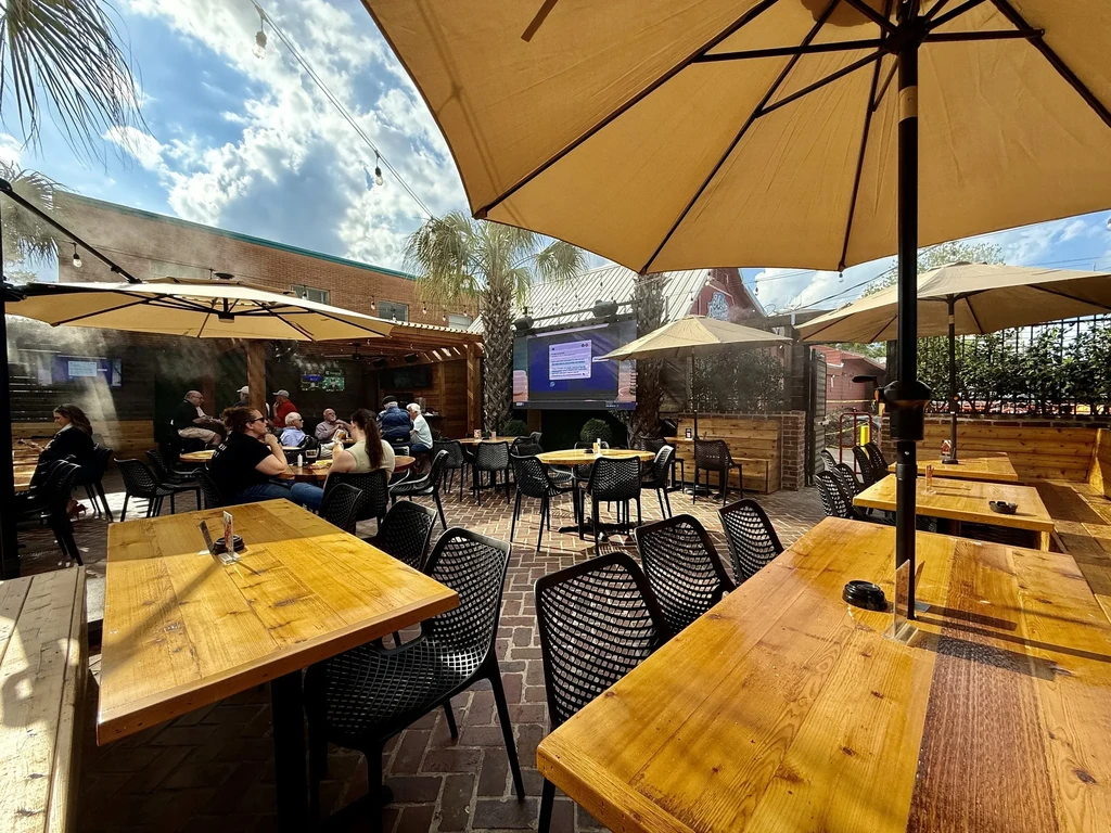 Outdoor restaurant patio with tables, umbrellas, seated people, and large TV.
