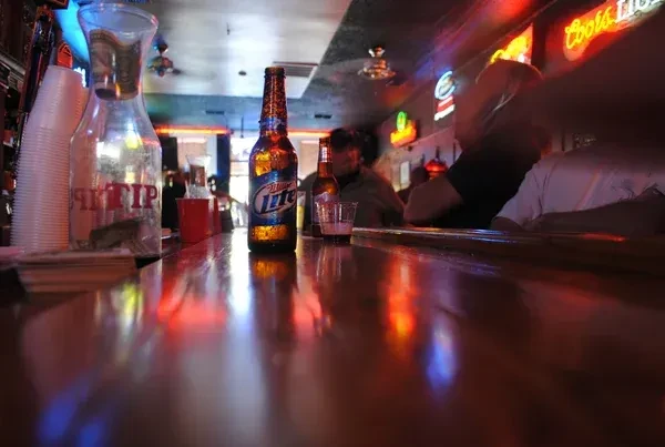 Low-angle bar view: frosty Miller Lite, tip jar, colorful neon reflections.