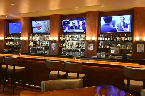 Modern bar interior with counter, stocked shelves, taps, and four TVs.