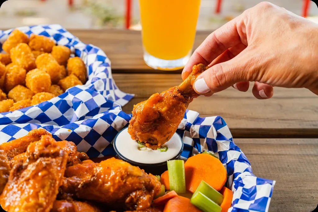 Hand dipping a buffalo chicken wing into dip, next to tater tots.