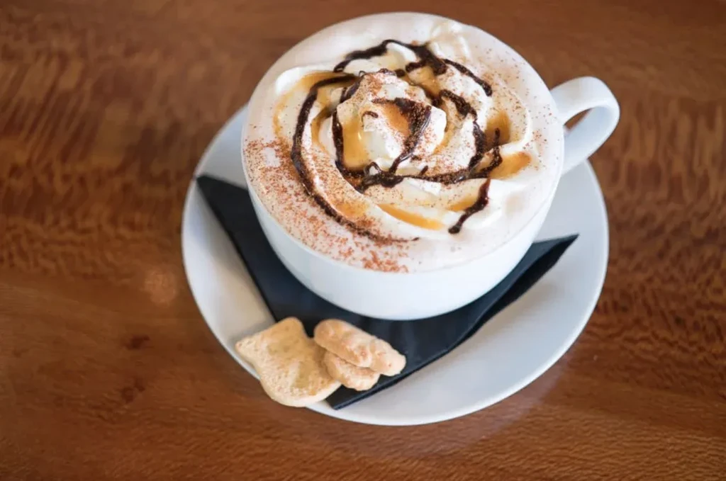 Hot drink with whipped cream, chocolate, caramel, and cookies on a wooden table.