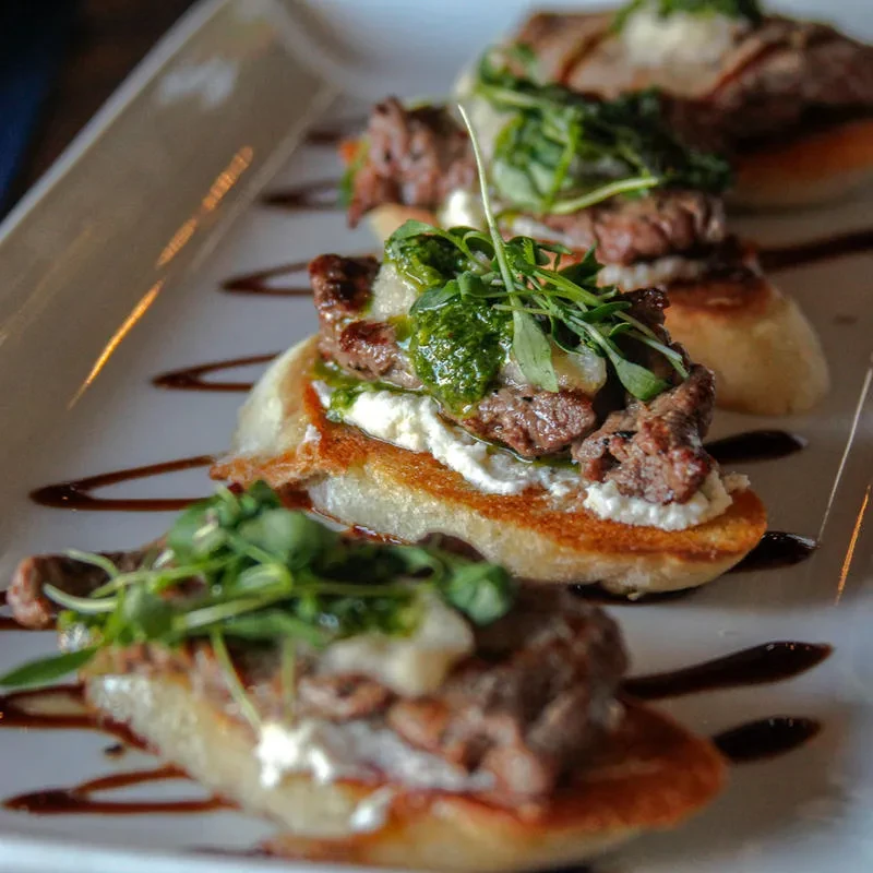 Plated crostini appetizer topped with meat, cheese, green sauce, and microgreens.