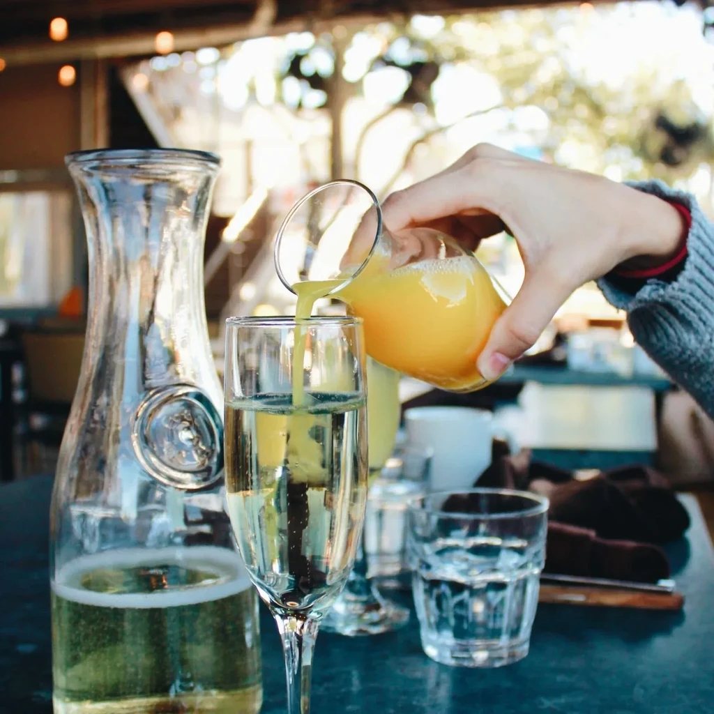 Hand pouring orange juice into a flute, making a Mimosa outdoors.