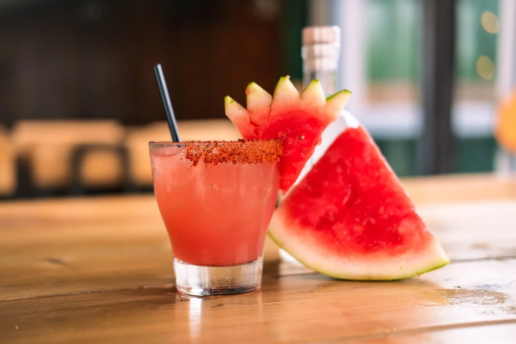 Red-orange cocktail with seasoned rim and watermelon slices on wood.
